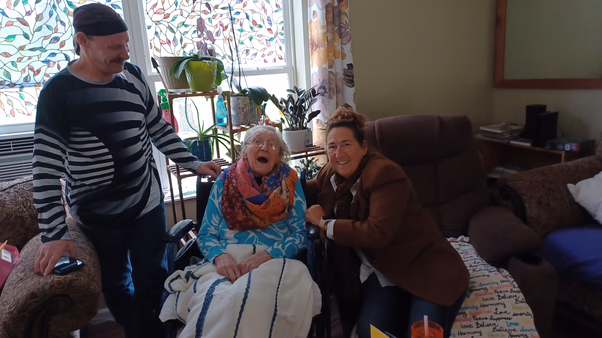 Three people in a cozy living room setting. An elderly woman in a wheelchair is in the center, wearing a blue patterned top and a colorful scarf, smiling and looking joyful. A man in a black and white striped sweater and a black cap stands to her left, resting his hand on the wheelchair. A woman in a brown jacket is seated to the right, leaning in close and smiling warmly. Behind them are windows with decorative leaf-patterned curtains and several potted plants on a stand. The room has comfortable furniture including a brown armchair and a couch with a blanket featuring words like 'Peace' and 'Love'.