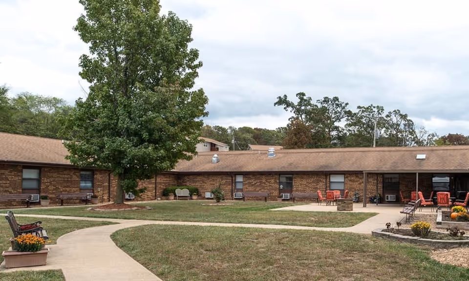 Courtyard of a single-story brick nursing center with a large tree, walkways, benches, and patio seating.