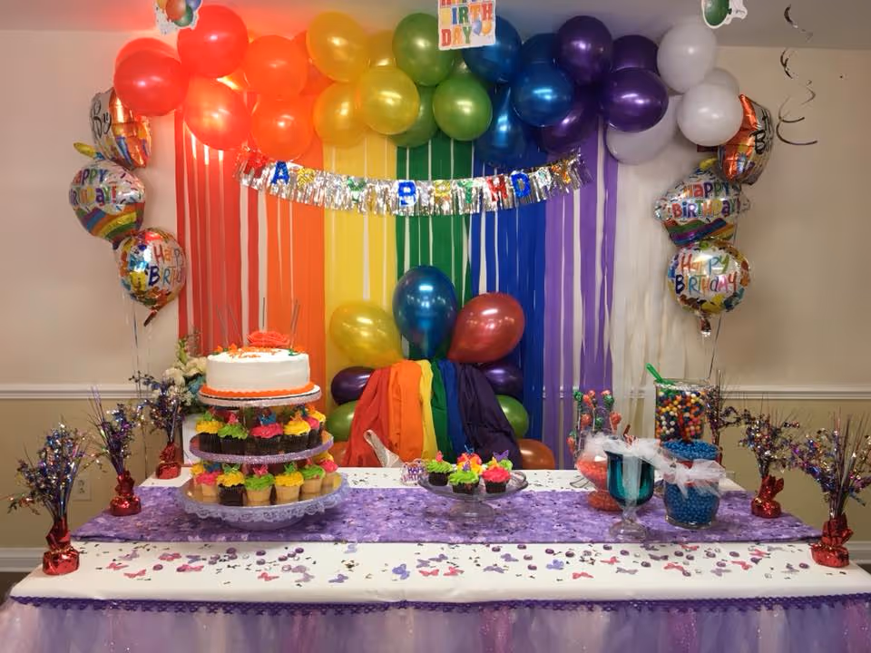 A colorful birthday party setup with a rainbow balloon arch and matching rainbow streamers on the wall. A table is decorated with a purple runner, confetti, and a tiered stand holding a white birthday cake with orange trim and multiple cupcakes with bright frosting. There are also jars filled with colorful candies and several 'Happy Birthday' balloons on either side of the table.