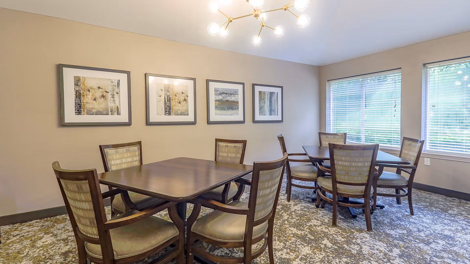 A well-lit dining room with two wooden tables, each surrounded by six cushioned chairs. The room has beige walls adorned with four framed abstract paintings. Large windows with blinds allow natural light to fill the space, and a modern chandelier hangs from the ceiling. The floor is covered with a patterned carpet.