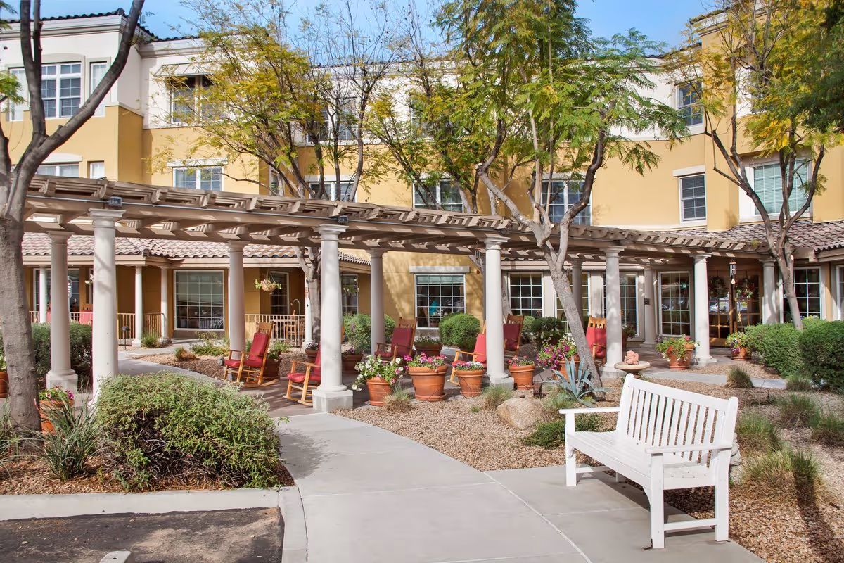 Courtyard in front of a multi-story senior living building with a pergola, potted plants, rocking chairs and a white bench.