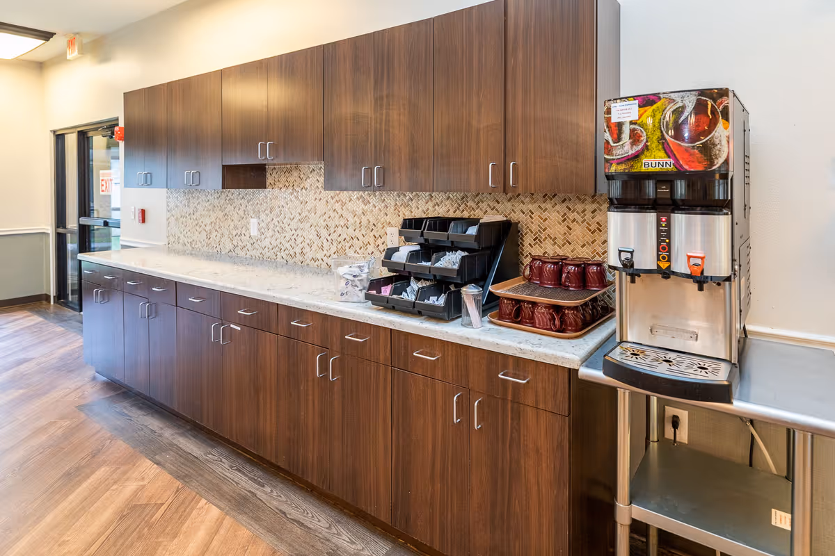 A long countertop with dark wooden cabinets above and below, a coffee machine with two dispensers on the right, and trays with cups and coffee supplies on the counter. The floor is wood-patterned, and there is a door with an exit sign in the background.
