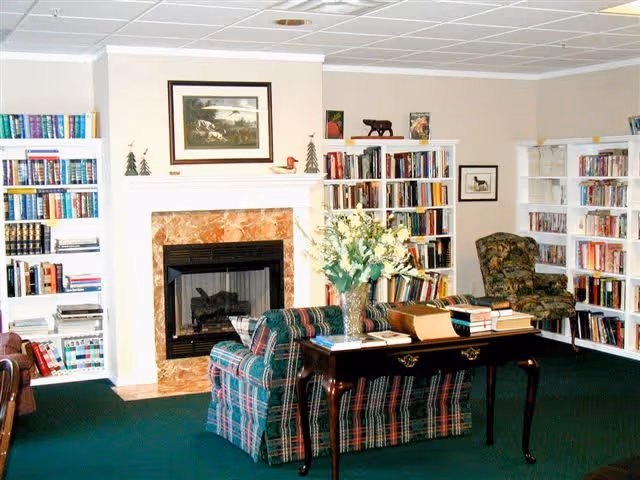 A cozy living room area with a green carpet, plaid upholstered sofa, and a wooden table with books and a vase of flowers. The room features a fireplace with a marble surround and a framed painting above it. Bookshelves filled with books line the walls, and there is a patterned armchair in the corner.
