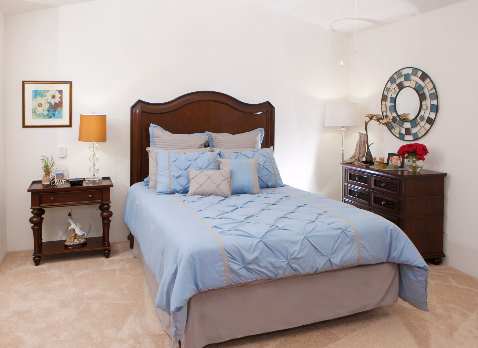 A neatly made bed with a wooden headboard, covered with light blue bedding and multiple pillows. On the left side of the bed is a wooden nightstand with a lamp, a small plant, and decorative items. On the right side is a wooden dresser with a round decorative mirror above it, a lamp, a vase with red flowers, and other small decor pieces. The room has beige carpet and white walls.