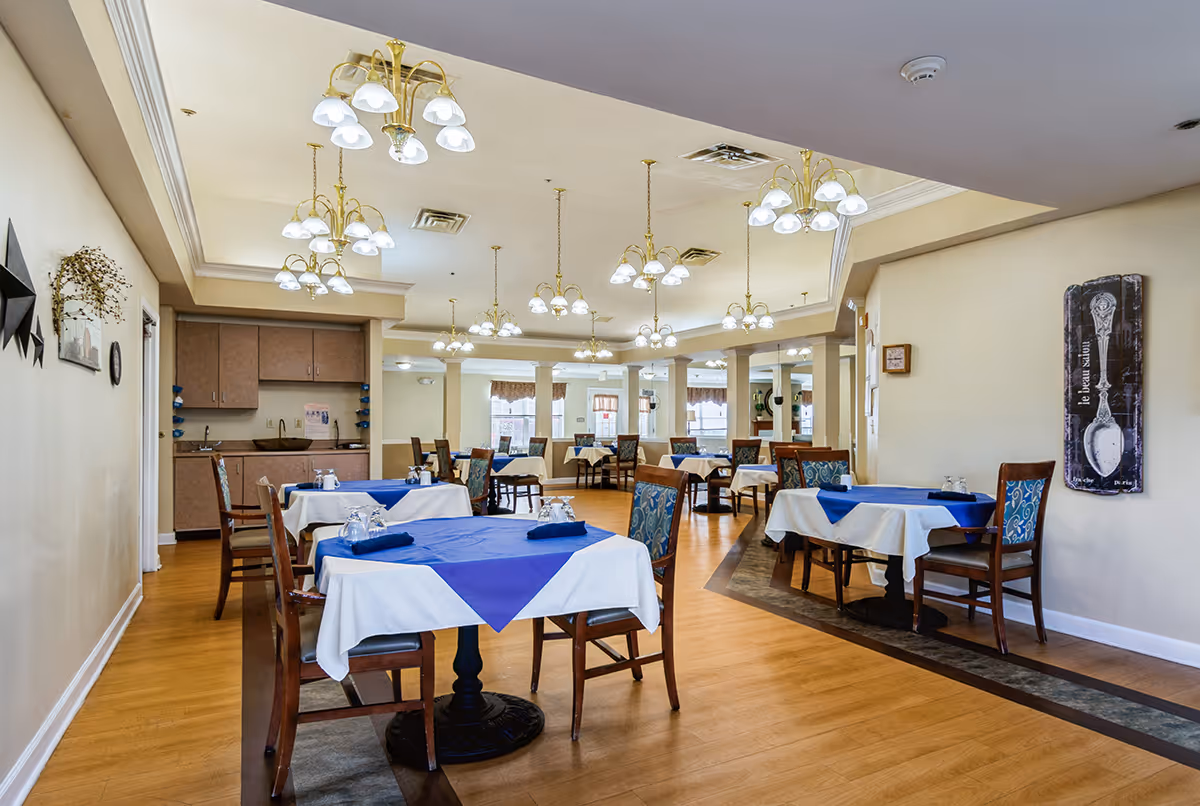 A spacious dining room with multiple tables covered in white tablecloths and blue table runners. Each table is set with glasses and napkins, surrounded by wooden chairs with patterned cushions. The room features wooden flooring, beige walls, multiple chandeliers with white lampshades hanging from the ceiling, and a small kitchenette area in the background.
