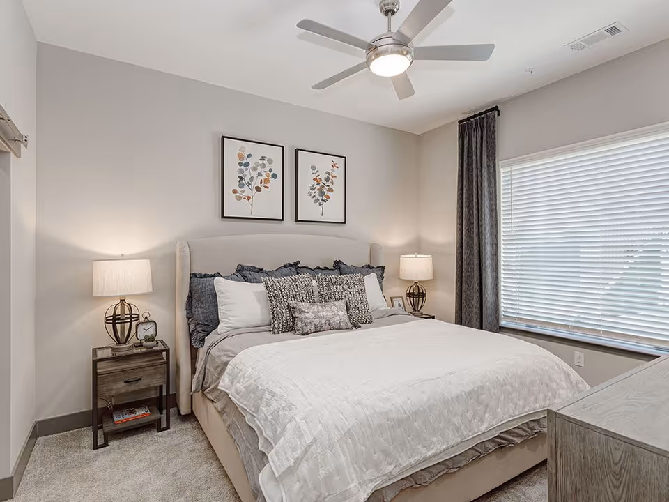 A neatly made bed with white and gray bedding in a bedroom. There are two framed botanical prints hanging above the bed. Two bedside tables each hold a lamp with a beige shade. A large window with closed blinds and dark curtains is on the right wall. A ceiling fan with a light is mounted on the ceiling.