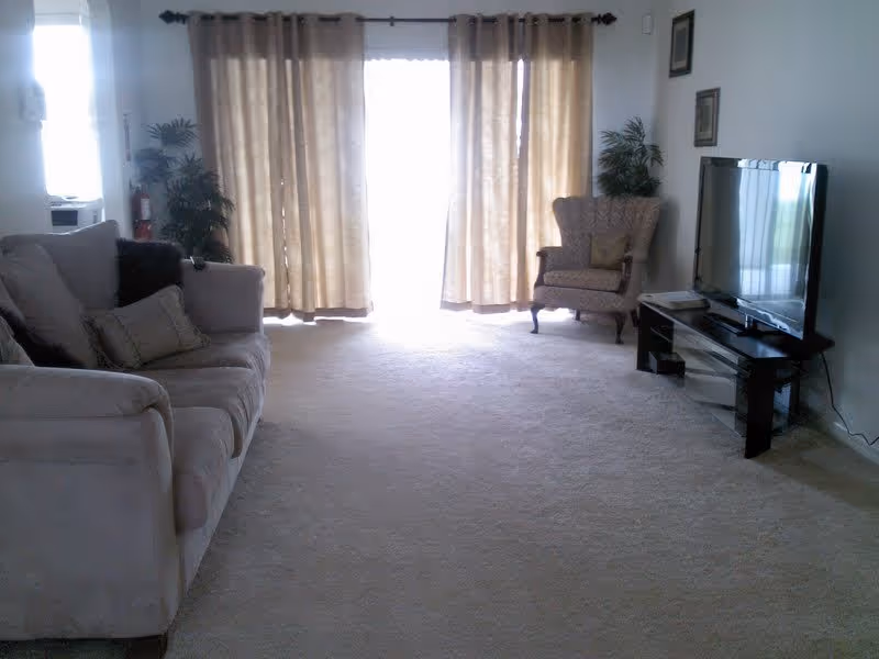 Bright living room with a beige sofa on the left, an armchair and flat-screen TV on the right, and large curtained sliding doors letting in light.