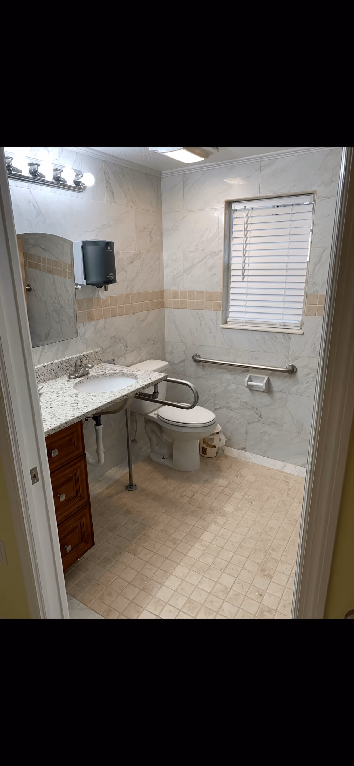 A clean bathroom with beige tiled floor and marble-patterned walls. It features a white toilet with a metal grab bar on the side and behind it, a granite countertop with an under-mount sink and a faucet. Above the sink is a mirror and a wall-mounted paper towel dispenser. A window with white blinds is on the wall above the toilet, and there is a toilet paper holder mounted on the wall. The bathroom has bright overhead lighting.