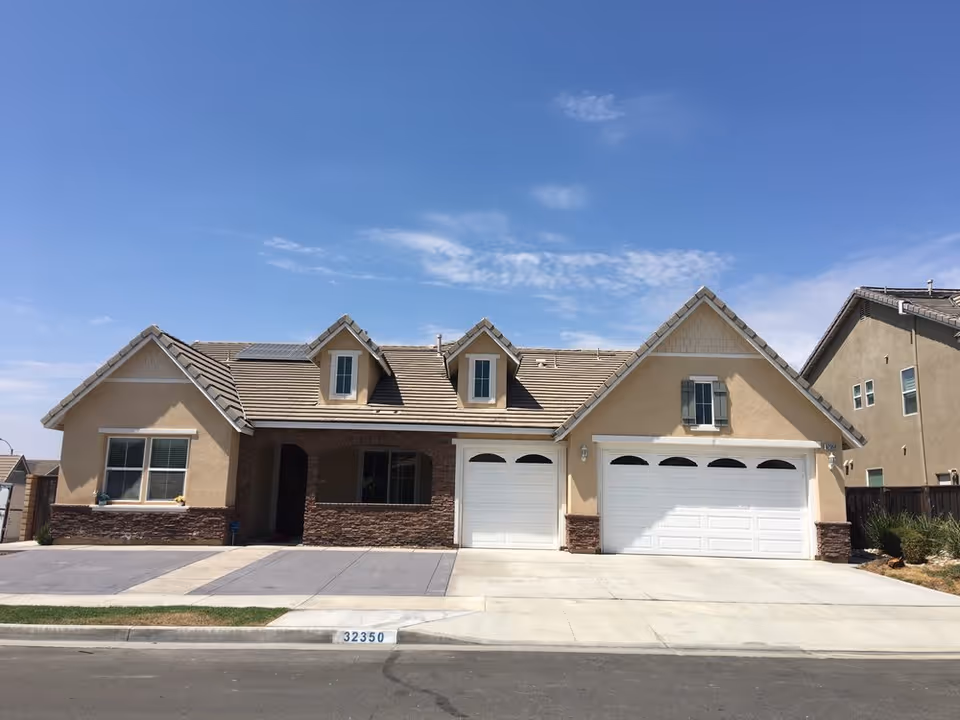 Single-story suburban house with a three-car garage, dormer windows, and a front driveway under a clear blue sky.