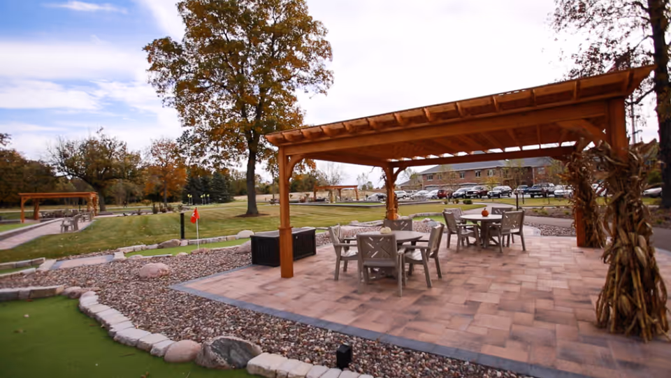 Outdoor patio area with a wooden pergola covering several tables and chairs, surrounded by landscaped grounds with trees, a putting green, and a building with parked cars in the background under a partly cloudy sky.