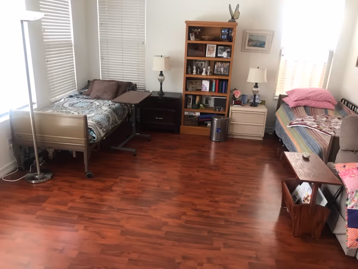 A room with two single beds, each with pillows and blankets. Between the beds are two nightstands with lamps and a wooden bookshelf filled with framed photos and decorative items. The room has wooden flooring and white walls with windows covered by blinds.