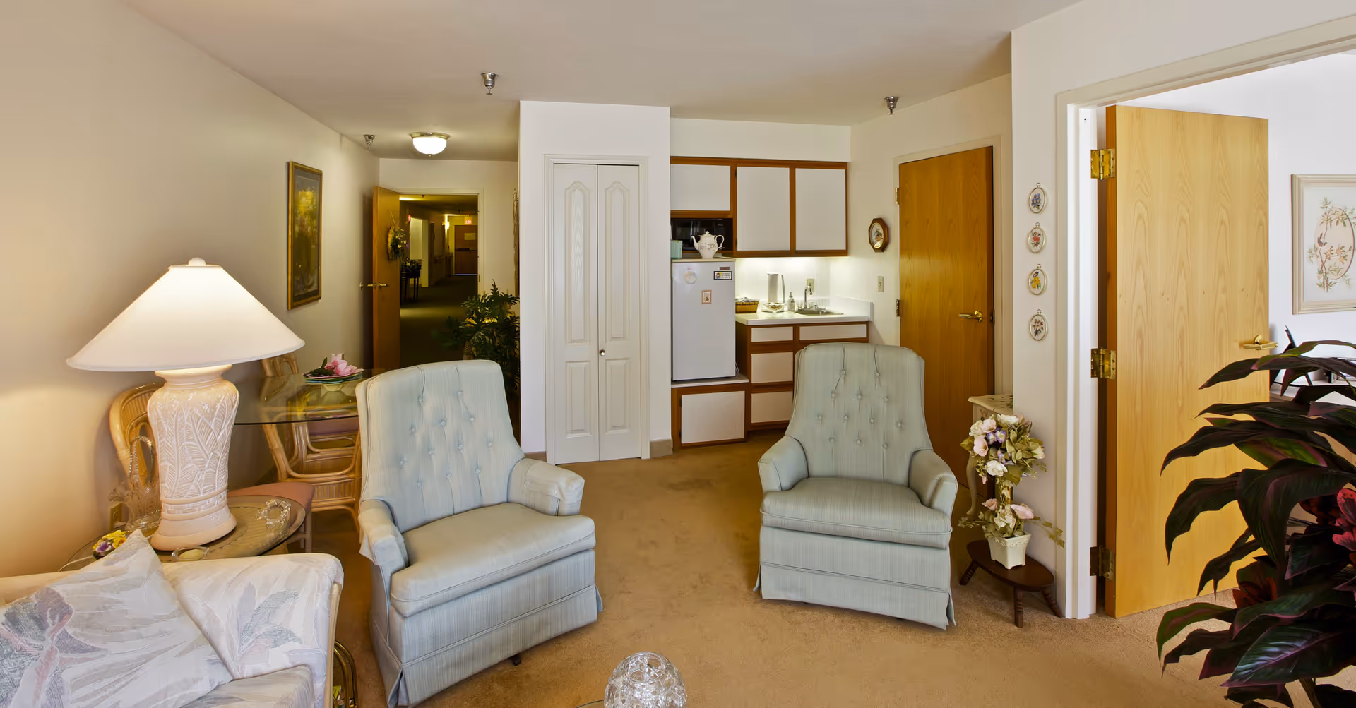 Interior view of a senior living facility apartment showing a small living area with two light blue armchairs, a glass side table with a lamp, and a small kitchenette in the background. There is a hallway leading to other rooms, a wooden door open to another room, and some decorative plants and wall art.