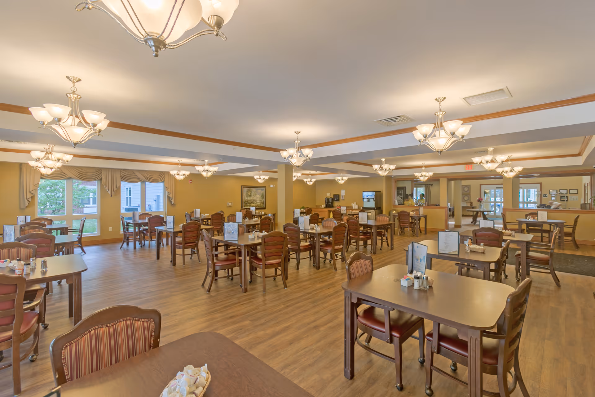A spacious dining room with multiple wooden tables and chairs arranged neatly. The room has large windows with beige curtains allowing natural light to enter. Several chandeliers hang from the ceiling, illuminating the space. Menus and condiments are placed on the tables, and the floor is covered with wood-style flooring.