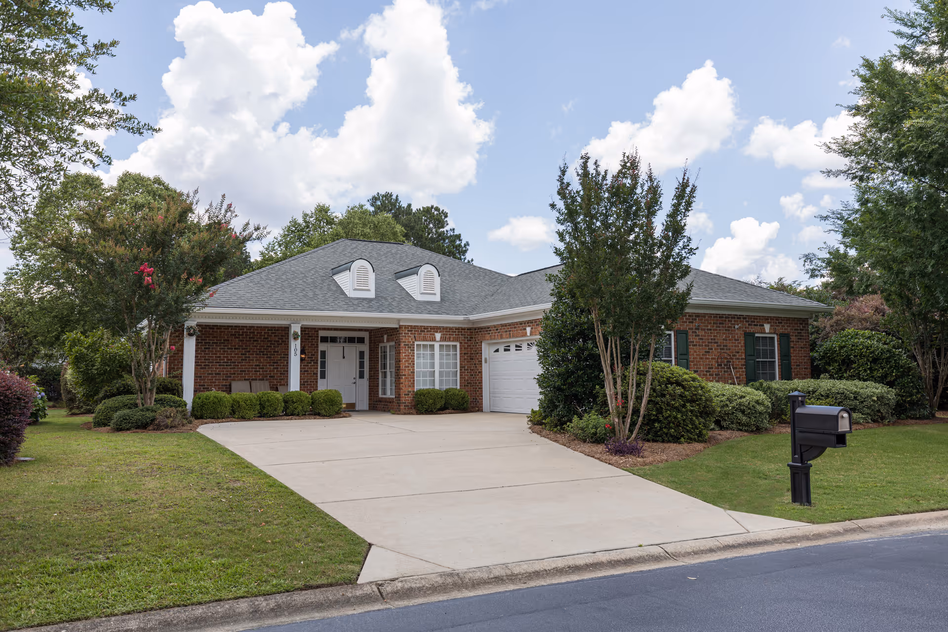 Front view of a single-story brick house with a driveway, garage, and mailbox surrounded by landscaping under a partly cloudy sky.