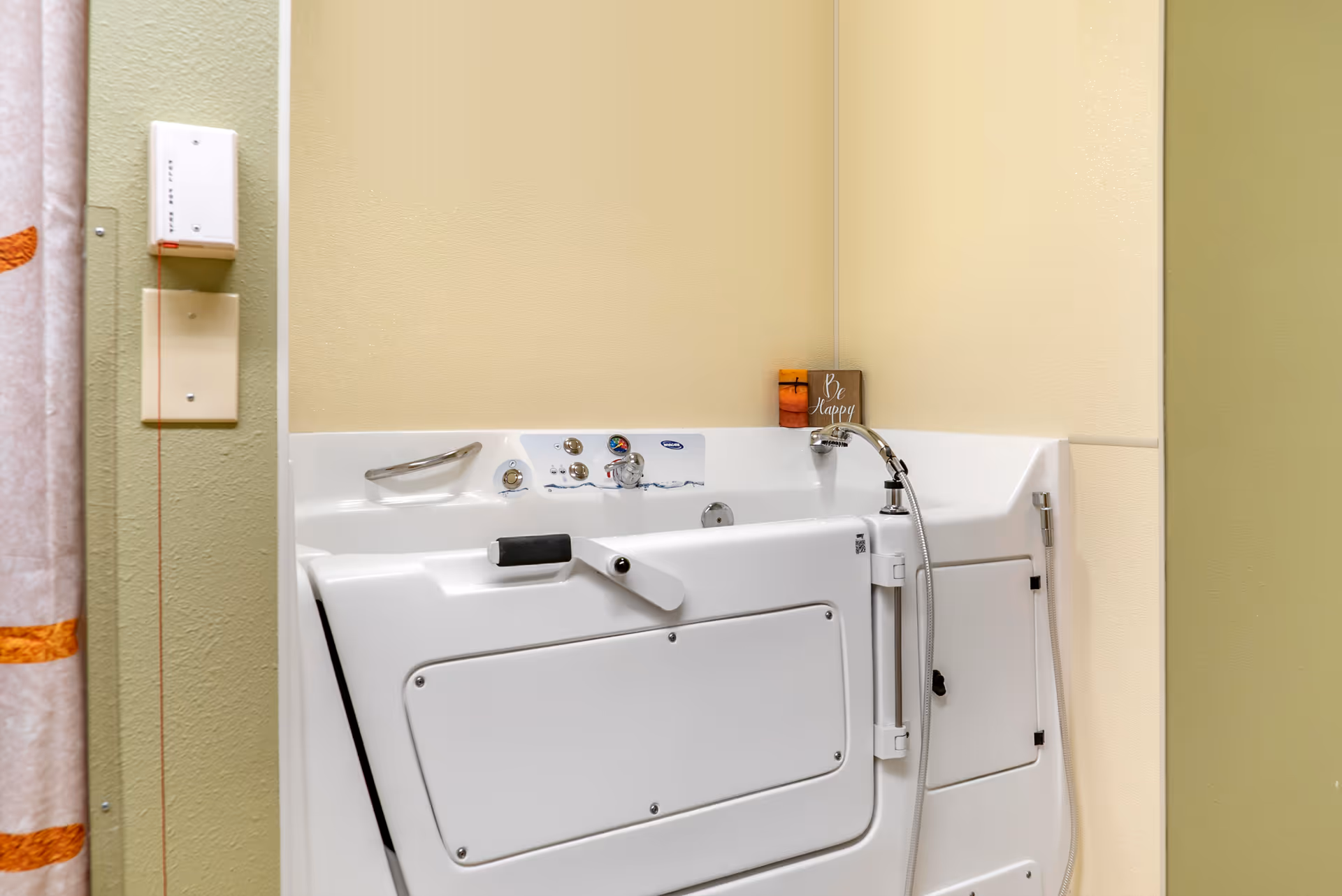 A white walk-in bathtub with a door and handrails installed in a bathroom corner with beige walls. There is a shower curtain with orange stripes partially visible on the left side and a small decorative sign that says 'Be Happy' placed on the bathtub ledge.