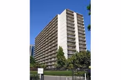 Tall multi-story residential building with balconies on each floor, surrounded by trees and a fenced area under a clear blue sky.