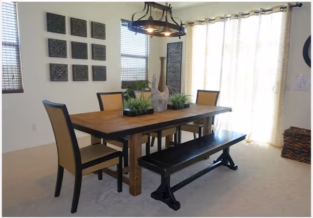A dining room with a wooden table surrounded by two chairs with beige cushions and a black bench. The table has decorative plants and a wooden sculpture. The room features large windows with blinds and light-colored curtains, and wall art consisting of square panels.