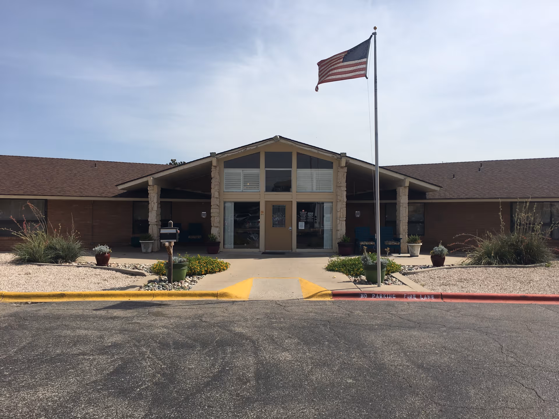 Front entrance of a single-story nursing facility with a flagpole flying an American flag and low landscaping.