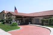 Front entrance of a single-story senior living building with a covered porte-cochère, American flag, and curved brick driveway.