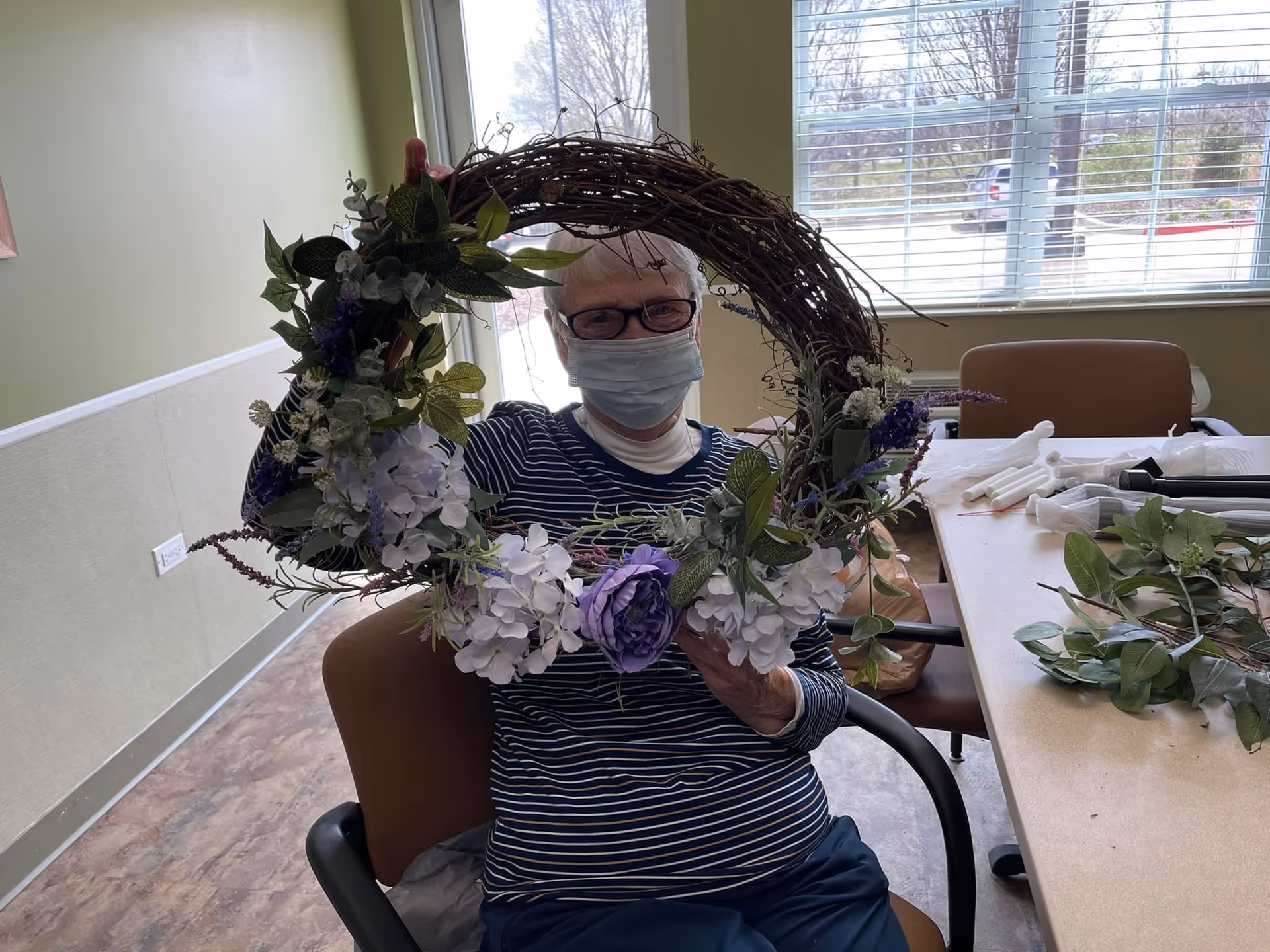 An elderly woman wearing glasses and a face mask is sitting in a chair inside a room with light green walls and a window with blinds. She is holding up a large decorative wreath adorned with purple and white flowers and green leaves. On the table next to her are various craft supplies and greenery.