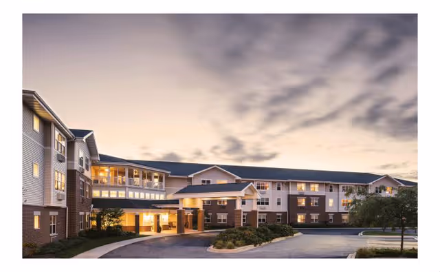 Front exterior of a multi-story senior living building with a covered entrance and illuminated windows at dusk.