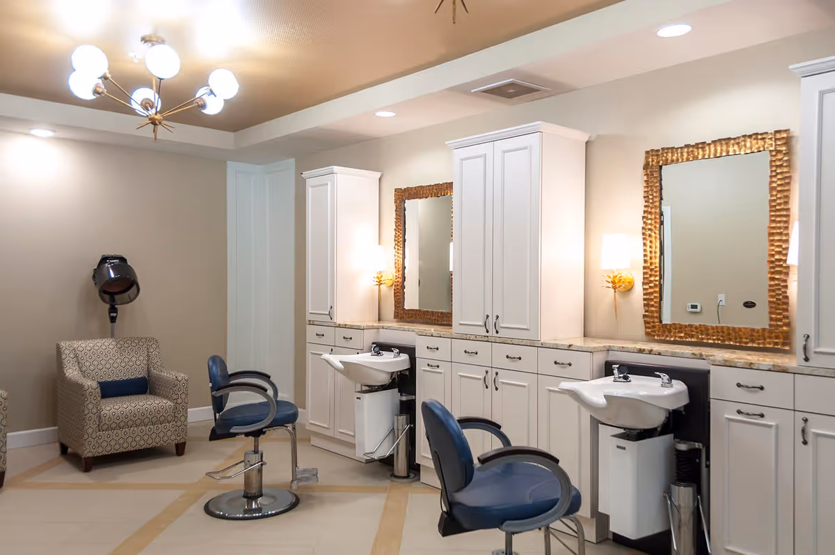 Interior view of a salon area with two blue salon chairs in front of white cabinetry and sinks for hair washing. Two large mirrors with decorative gold frames are mounted on the wall above the sinks. There is a patterned armchair and a hair dryer hood in the corner. The room has beige walls and a modern chandelier on the ceiling.