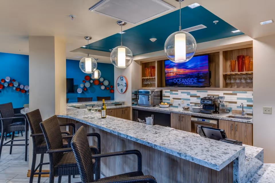 Communal bar-style kitchenette with marble countertops, wicker bar stools, pendant lights, coffee machines and a wall-mounted TV.