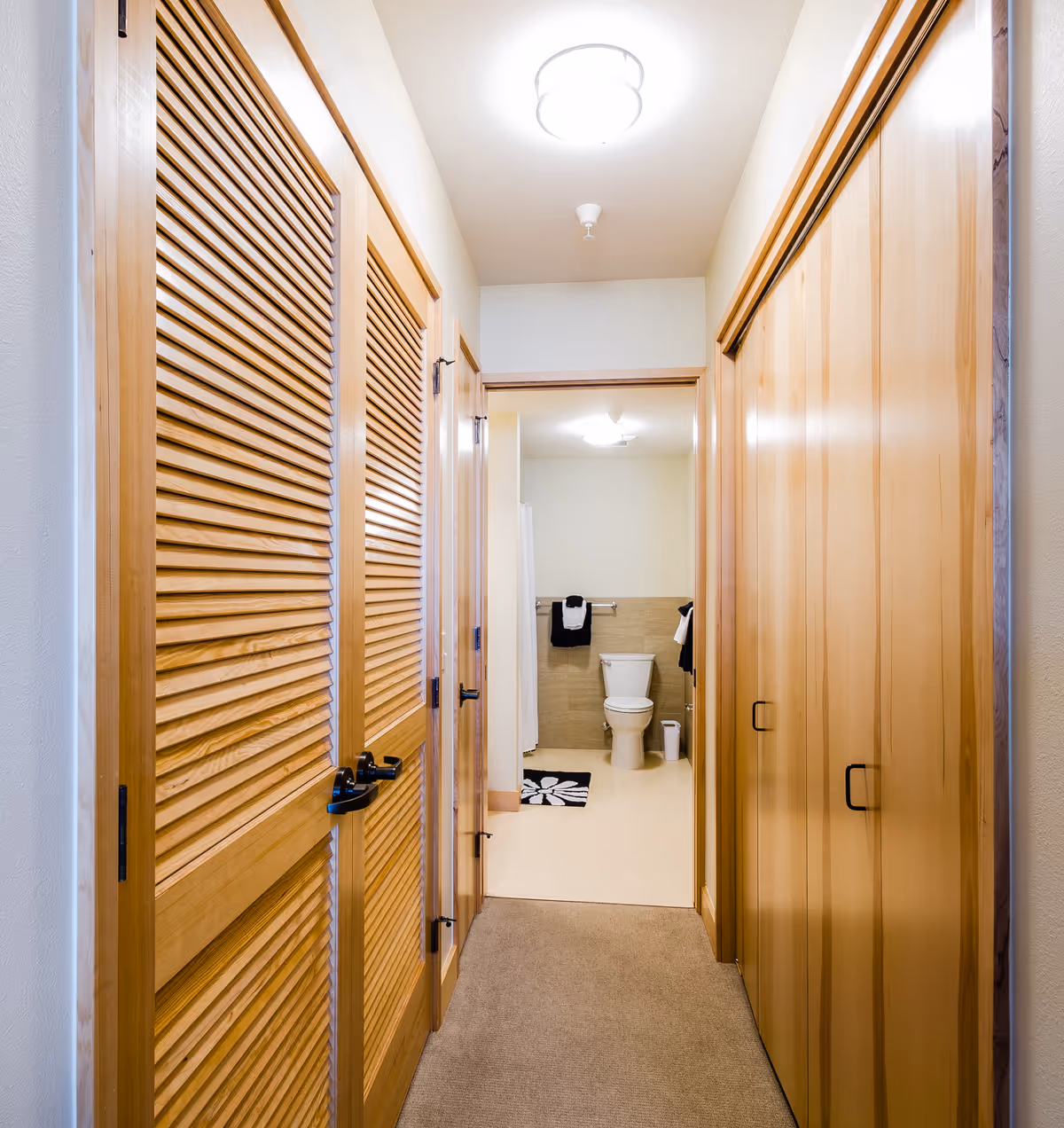 A hallway with wooden louvered doors on the left and wooden sliding closet doors on the right, leading to a bathroom with a toilet, towel rack with black and white towels, and a small trash can.