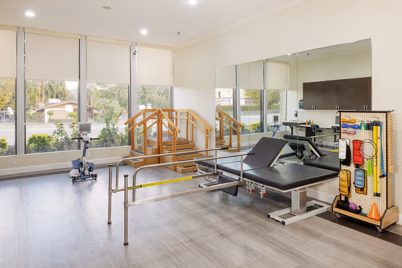 A physical therapy room with large windows letting in natural light. The room contains parallel bars for walking exercises, a padded adjustable therapy table, a wooden staircase with handrails for stair training, and a pegboard with various therapy tools and equipment. A large mirror covers one wall, reflecting the room.