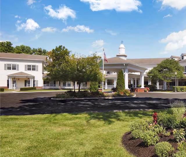 Front exterior view of The Cottage at Litchfield Hills senior living facility with a circular driveway, landscaped greenery, trees, and an American flag under a blue sky with scattered clouds.