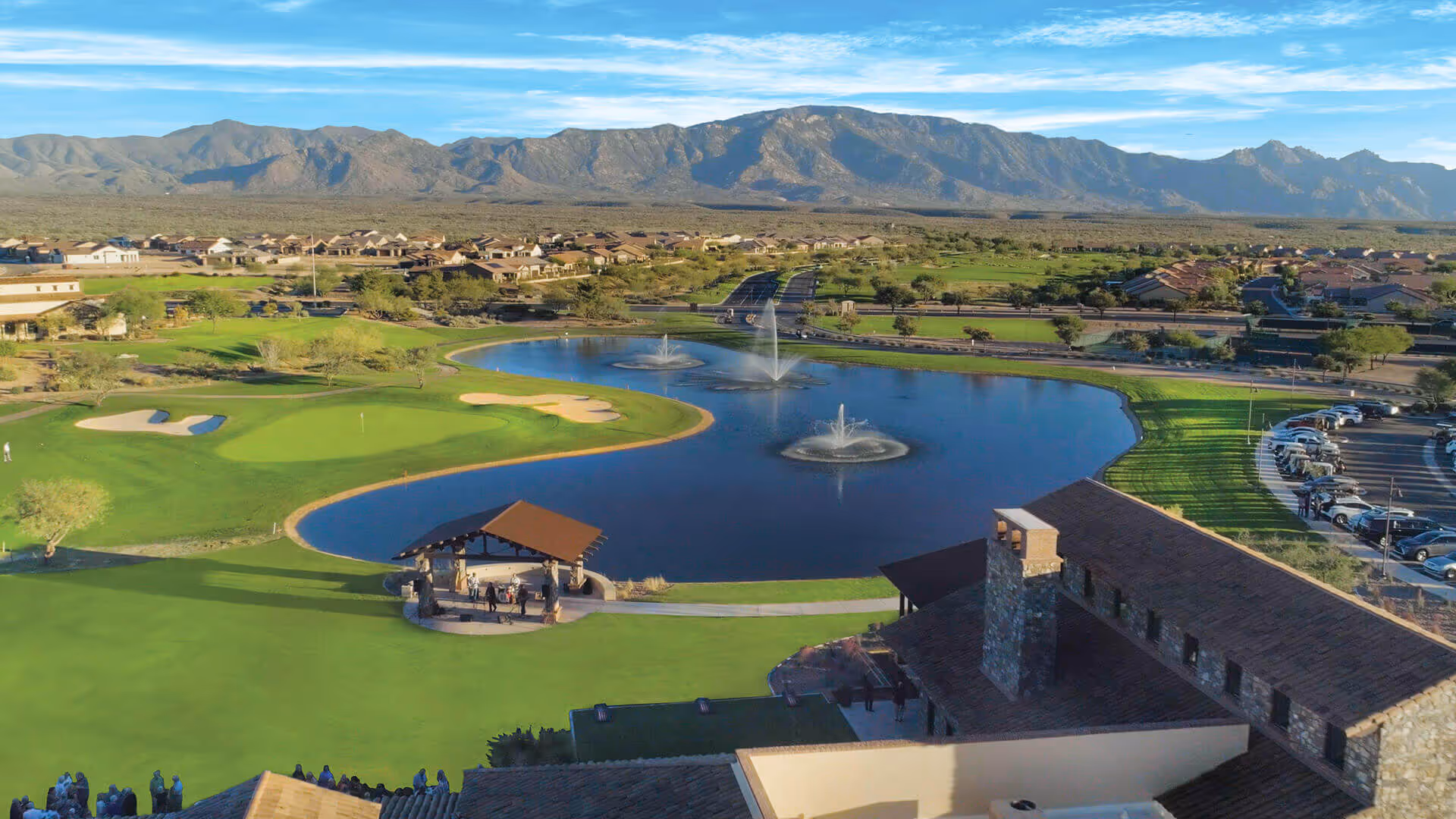 Aerial view of a senior living facility with a large pond featuring three water fountains, a golf green with sand traps, a covered pavilion with people underneath, surrounding green lawns, residential buildings in the background, a parking lot with cars, and mountains in the distance under a clear blue sky.