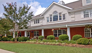 Exterior view of a two-story senior living facility building with a combination of brick and light-colored siding. The building features multiple windows, a well-maintained lawn, shrubs, and a tree under a partly cloudy sky.