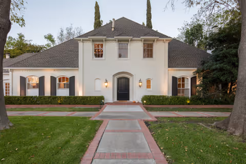Front exterior view of a large, white two-story house with a dark gray roof, black shutters, and a central black door flanked by two wall-mounted lanterns. The house is surrounded by green grass, trimmed hedges, and tall trees.