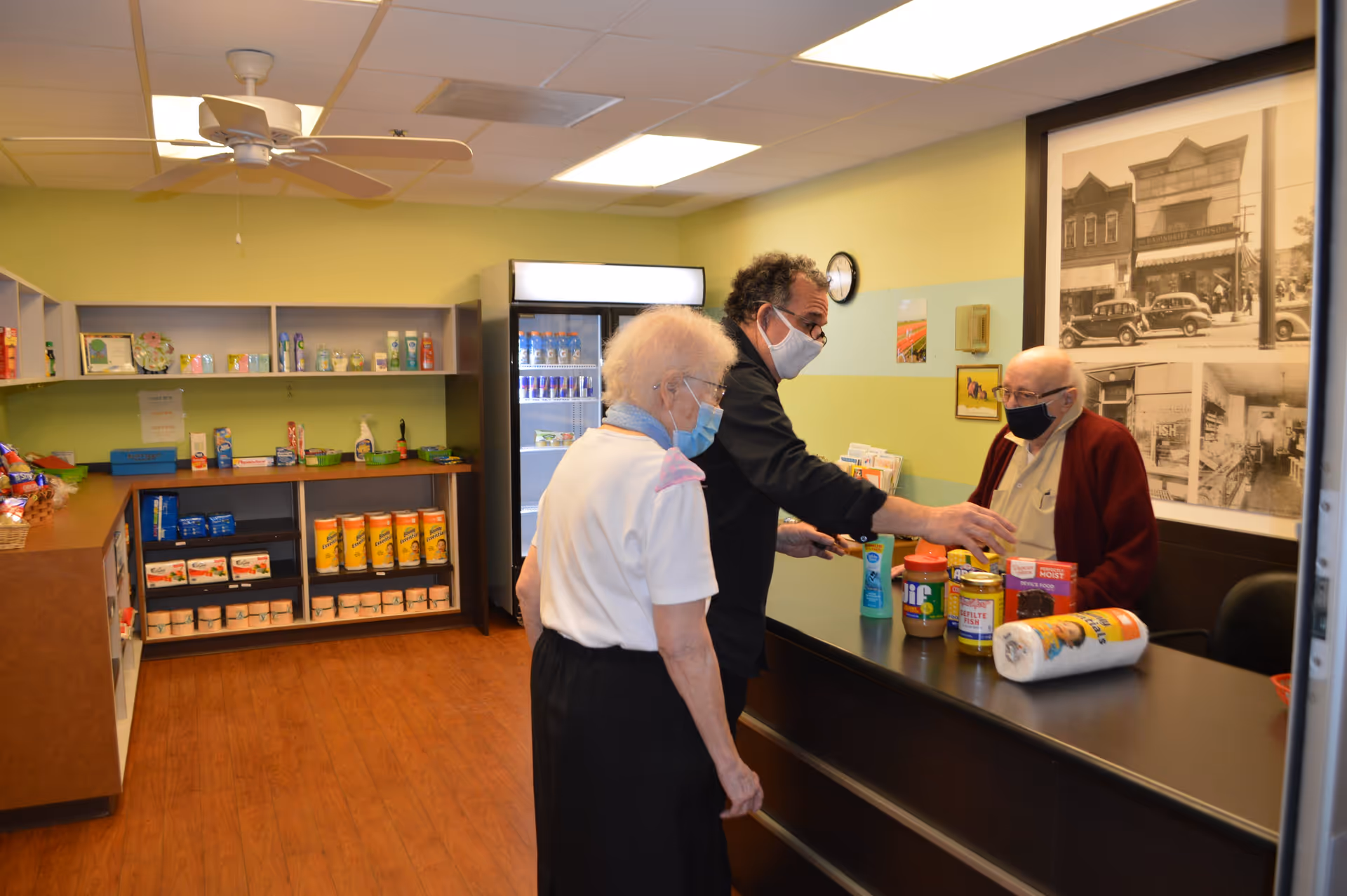 Three people wearing face masks inside a small store or pantry area with shelves stocked with various food and household items. One person is behind a counter with products like peanut butter, paper towels, and canned goods, while the other two stand on the opposite side of the counter. The room has light green walls, a ceiling fan, and a large black and white photo on the wall.