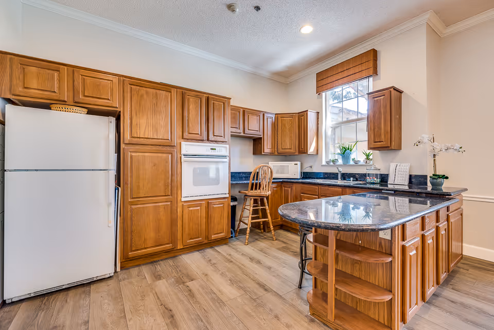 A bright kitchen with wooden cabinets, a white refrigerator, built-in oven, microwave, and a large granite countertop island with shelves. There is a window with a wooden blind above the sink, and several potted plants on the countertop and windowsill. The floor is light wood, and the walls are painted a light color.