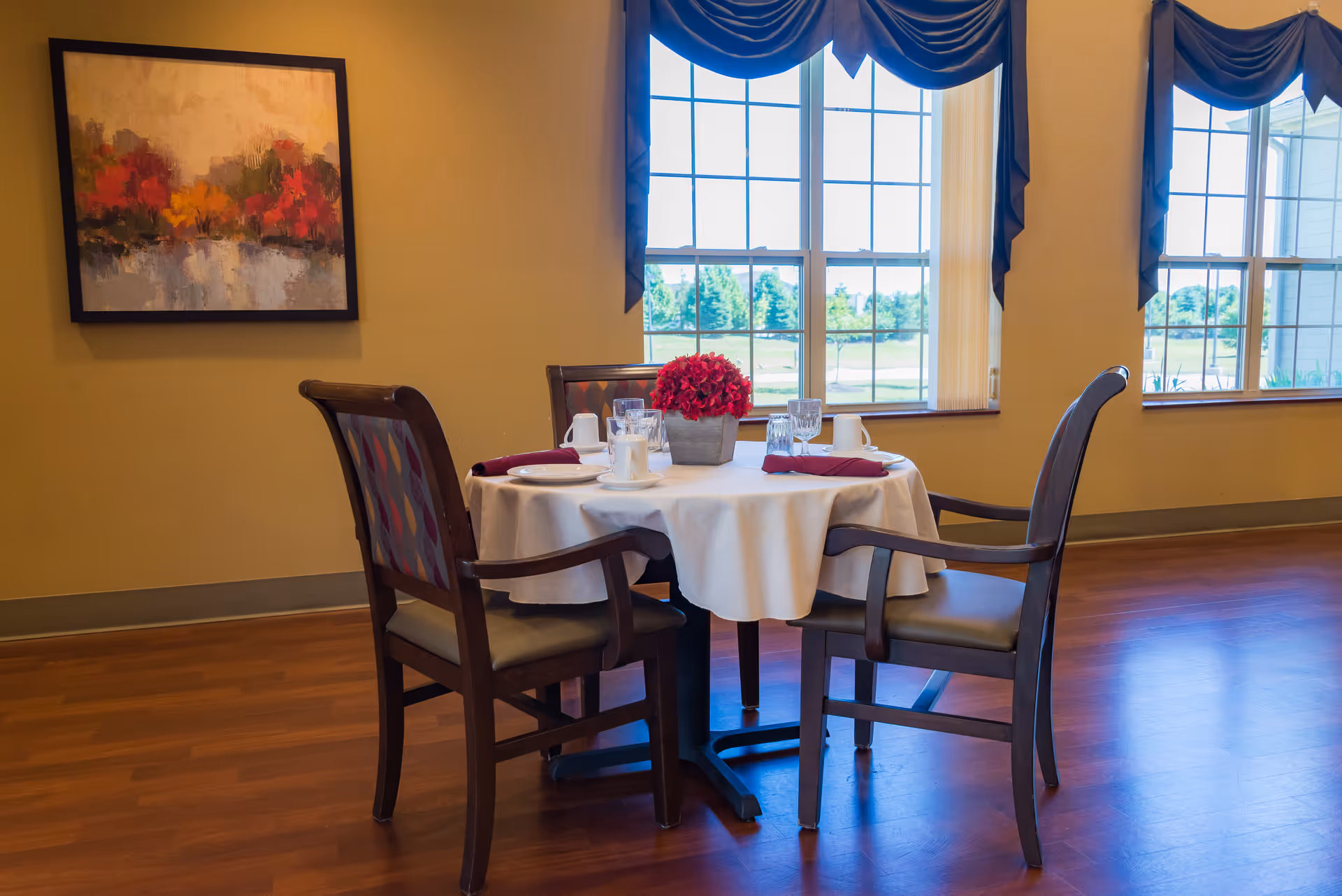 A round dining table set for four with a white tablecloth, red napkins, plates, cups, and glasses. A small pot of red flowers is in the center of the table. The room has wooden flooring, beige walls, two large windows with dark blue valances, and a colorful abstract painting on the wall.