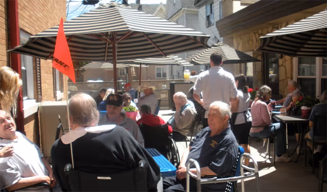 A group of elderly people sitting and socializing outdoors under large striped umbrellas in a courtyard area. Some individuals are in wheelchairs, and there are caregivers or staff members attending to them. The setting appears to be a communal outdoor space at a senior living facility.