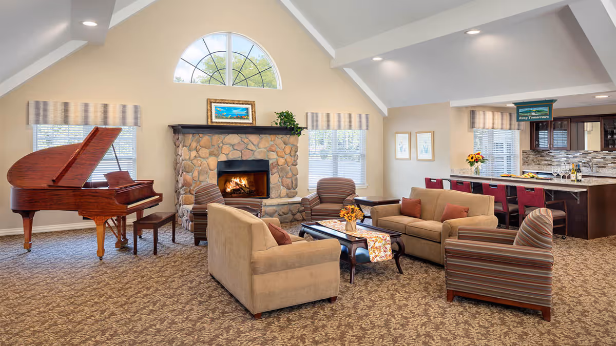 A spacious living room with a stone fireplace, a grand piano, and comfortable seating including two beige sofas and two striped armchairs arranged around a coffee table. The room has large windows with valances, a high vaulted ceiling with recessed lighting, and a kitchen area with a bar counter and red bar stools in the background.