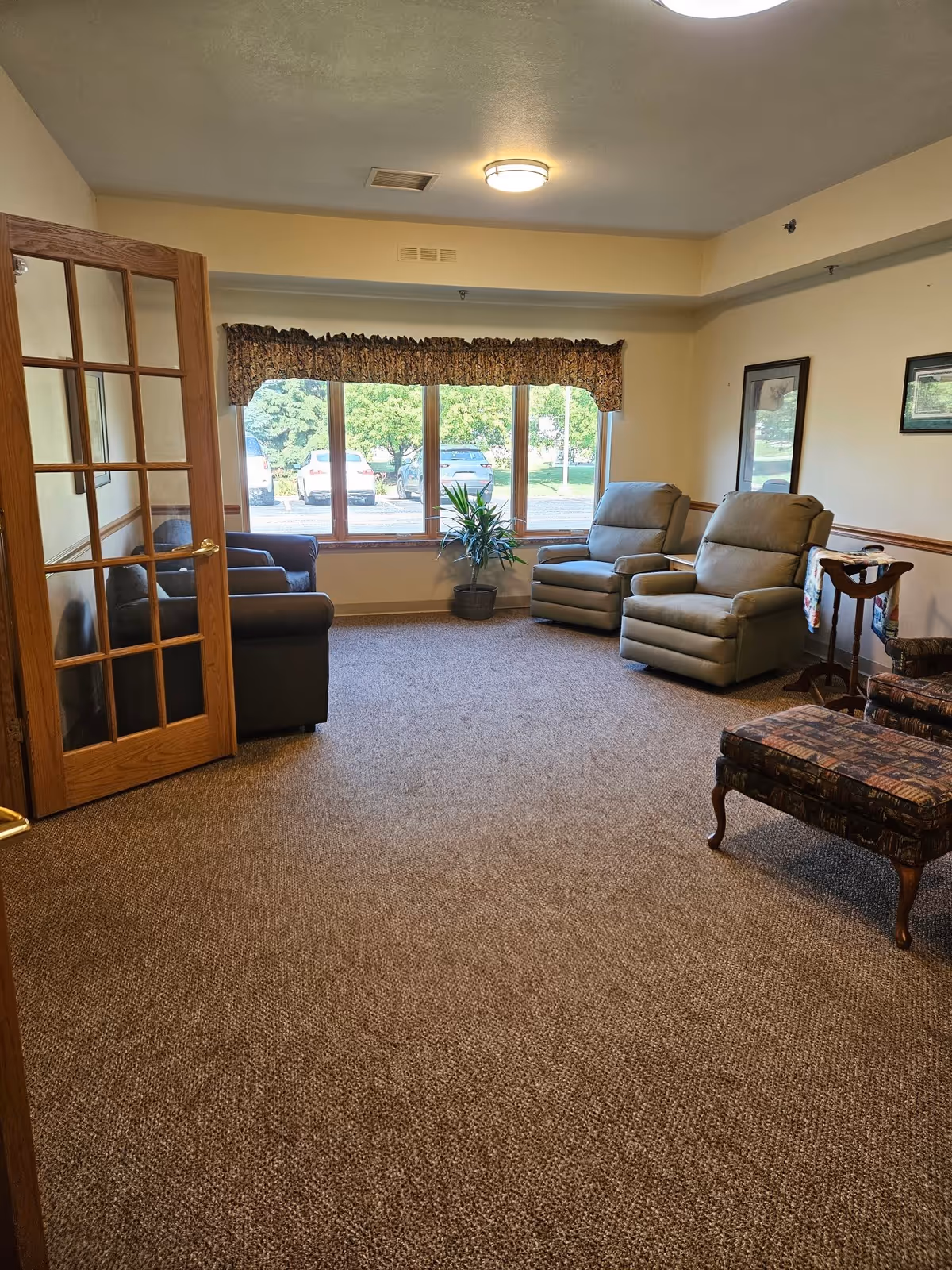 A cozy living room area with a large window showing parked cars and greenery outside. The room has a brown carpet, two beige recliner chairs, a dark brown couch, a patterned armchair with a matching ottoman, a small wooden side table with a decorative cloth, and framed pictures on the walls. A wooden door with glass panels is open on the left side.