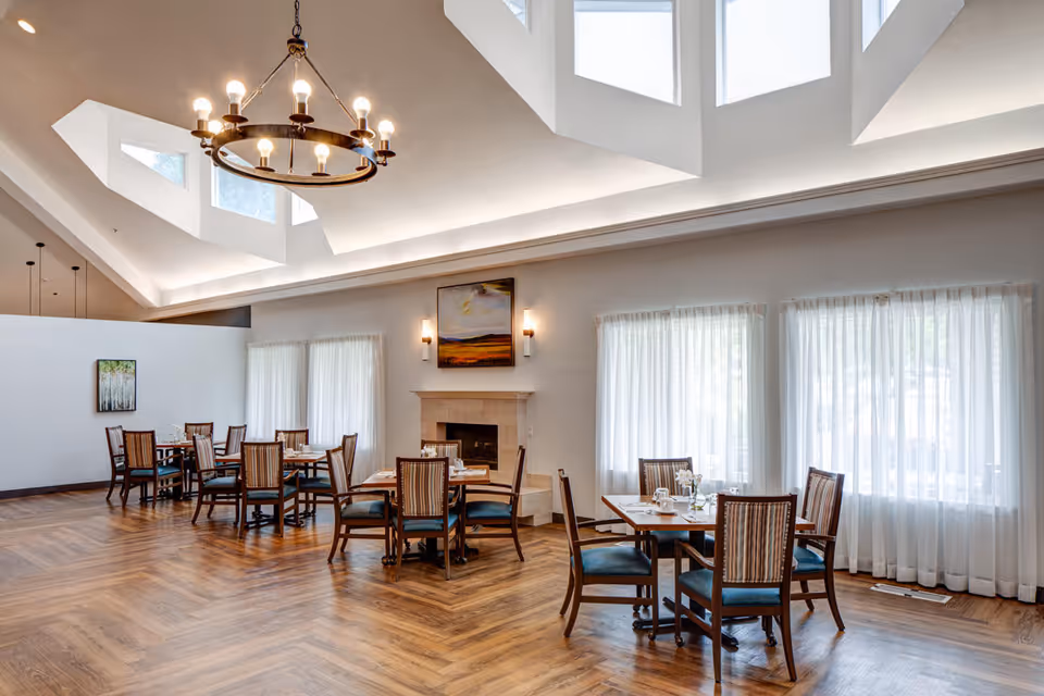 A bright dining room with wooden floors and several tables set with cups and plates. The room features large windows with sheer white curtains, a fireplace with a painting above it, and a modern chandelier hanging from a high ceiling with skylights.