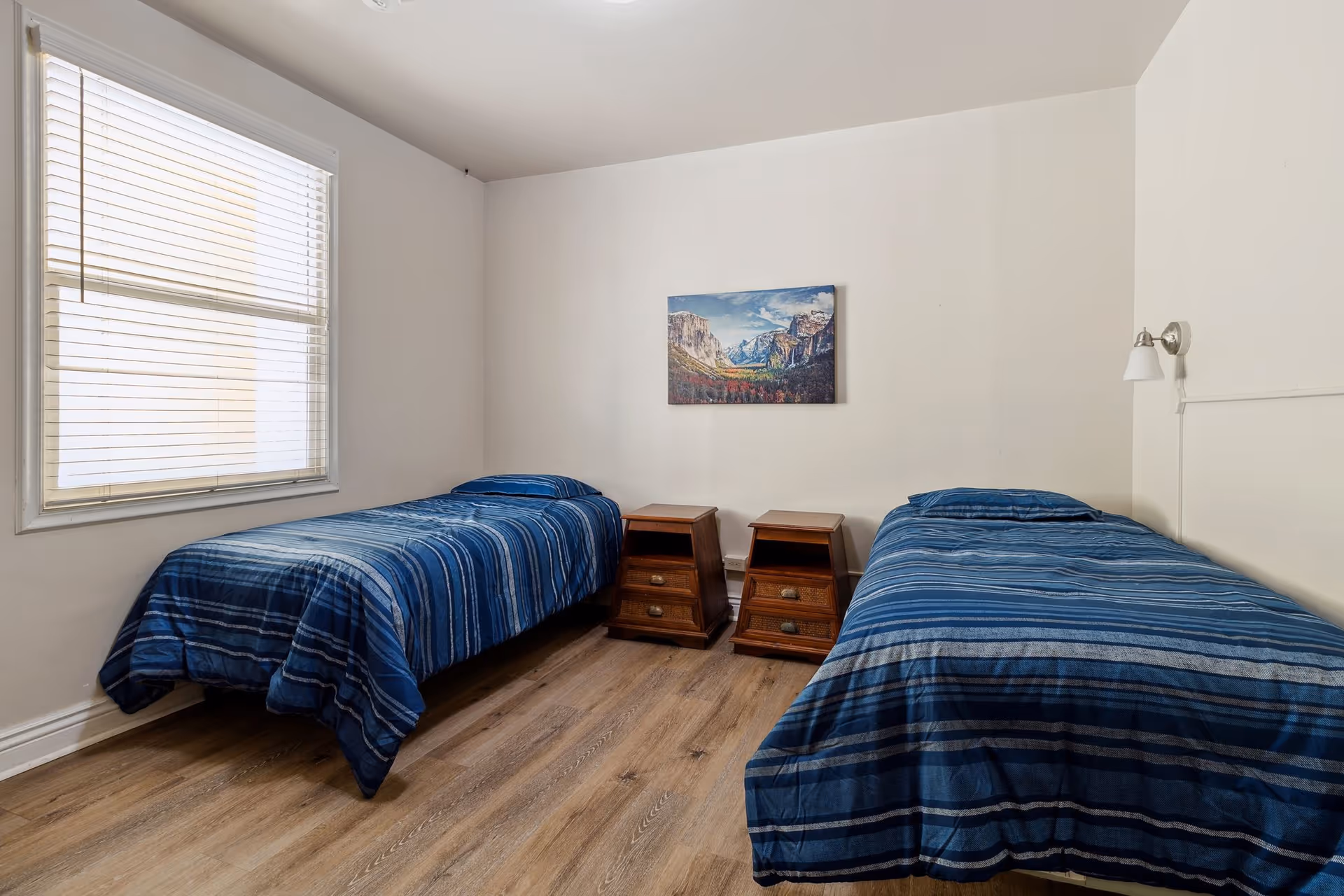 A simple bedroom with two single beds covered in blue striped bedding. Between the beds are two wooden nightstands with drawers. A window with blinds is on the left wall, and a landscape painting hangs on the wall above the nightstands. The room has light-colored walls and wood flooring.