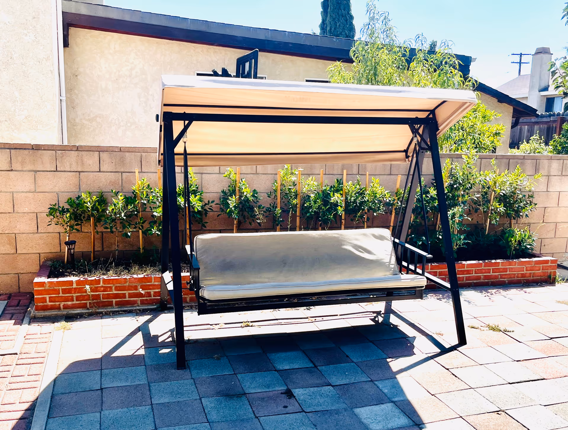 Outdoor patio area with a cushioned swing bench under a canopy, surrounded by a brick planter with green plants and a beige wall in the background.