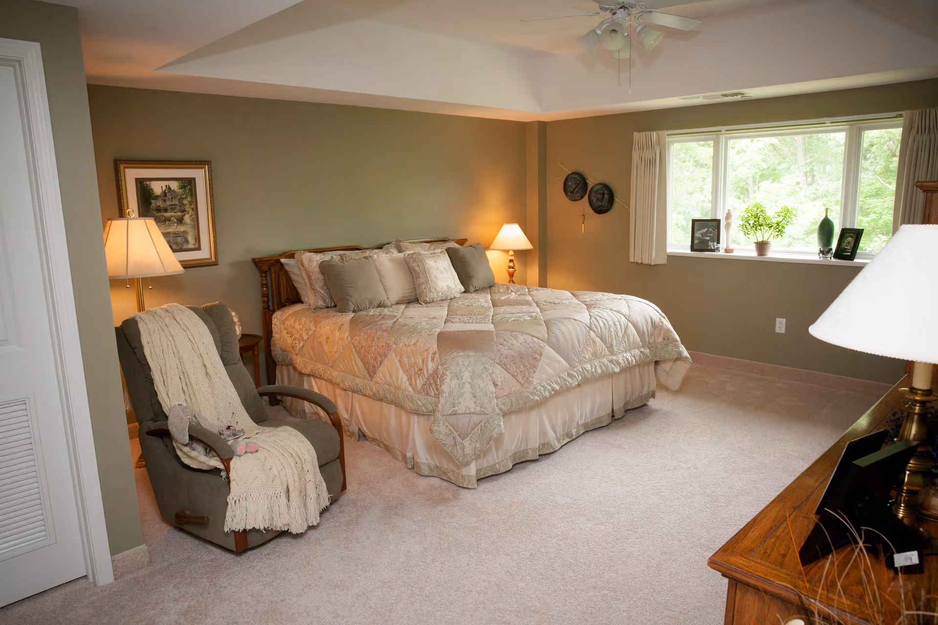 A cozy bedroom with a large bed covered in a beige quilt and multiple pillows. There is a comfortable armchair with a knitted throw and a small stuffed animal on it. Two bedside tables each have a lamp, and a framed picture hangs on the wall. A large window with light curtains lets in natural light, and a wooden dresser with a lamp and framed photos is visible in the foreground.