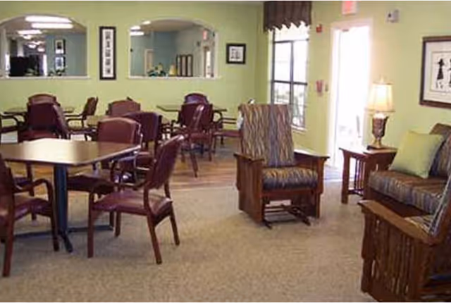 Bright communal room with several dining tables and chairs, upholstered armchairs, and soft green walls.