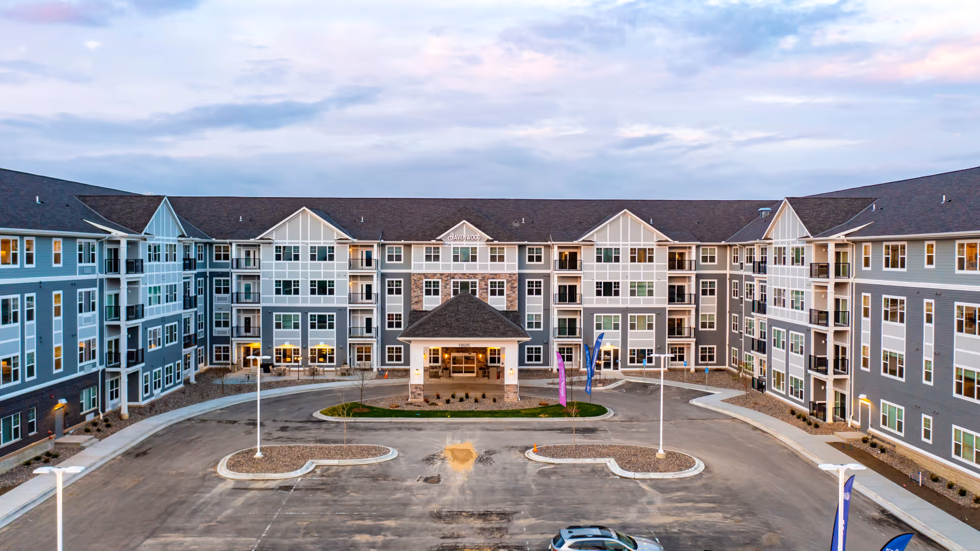 Front exterior view of Havenwood of Maple Grove, a large multi-story senior living facility with a covered entrance, multiple windows, balconies, and a spacious driveway with parking spaces and flag banners.