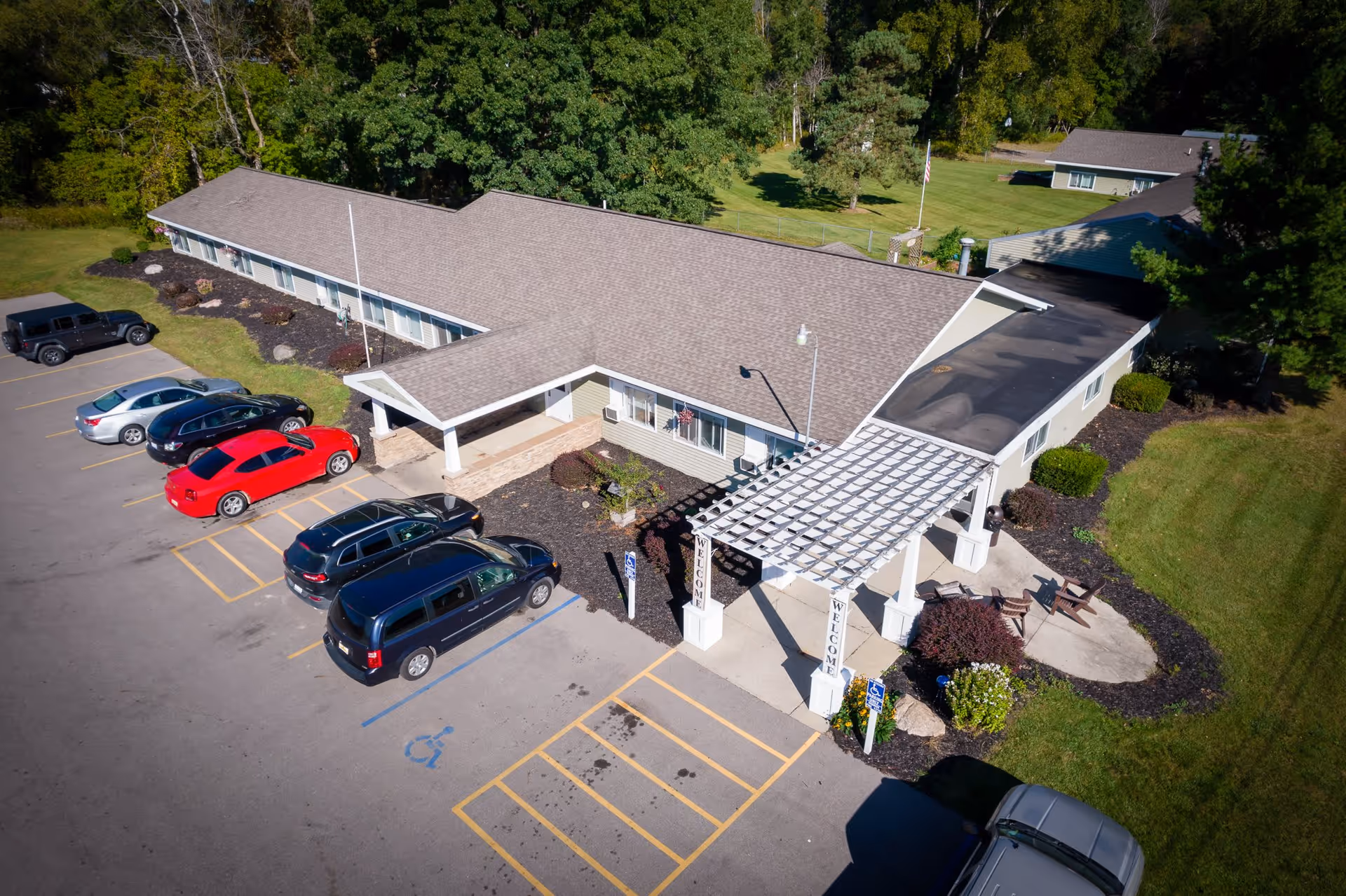 Aerial view of a single-story senior living facility building with a covered entrance and a pergola structure displaying 'WELCOME' signs. Several cars are parked in the parking lot, including handicapped spaces. The building is surrounded by green lawns and trees.