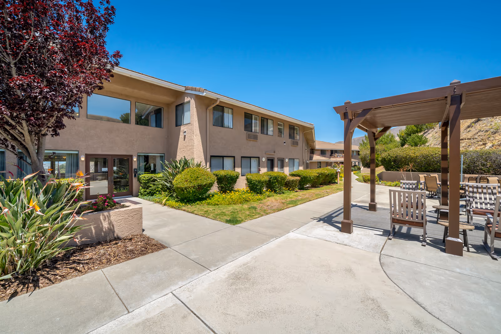 Outdoor view of a two-story beige building at Las Brisas Retirement Community with large windows and a landscaped garden featuring bushes and a tree with red leaves. A concrete walkway leads to the entrance, and there is a shaded pergola with chairs and tables on the right side under a clear blue sky.