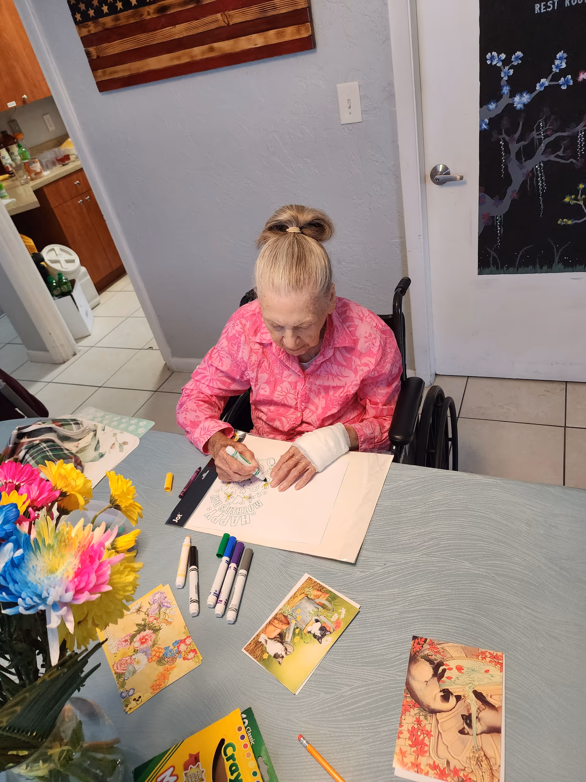 An elderly woman in a wheelchair wearing a pink floral shirt is coloring a picture at a table. The table has several markers, coloring pages, postcards, a pencil, and a box of Crayola markers. There is a colorful bouquet of flowers in a vase on the table. The room has tiled floors, a door with a decorative chalkboard, and a wooden American flag hanging on the wall.