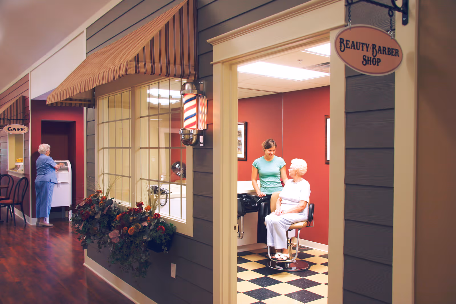 Interior hallway of a senior living facility showing a beauty/barber shop where a caregiver talks with an elderly woman in a salon chair and another resident near a cafe area.