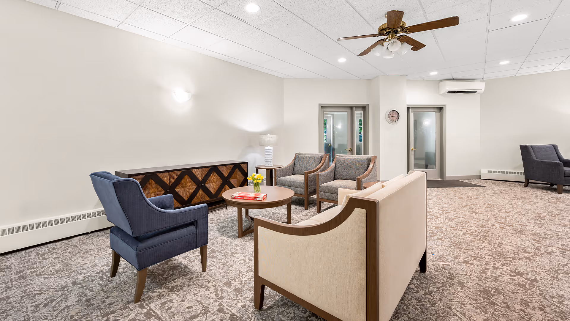 A well-lit lounge with sofas and armchairs arranged around coffee tables in a senior living facility.